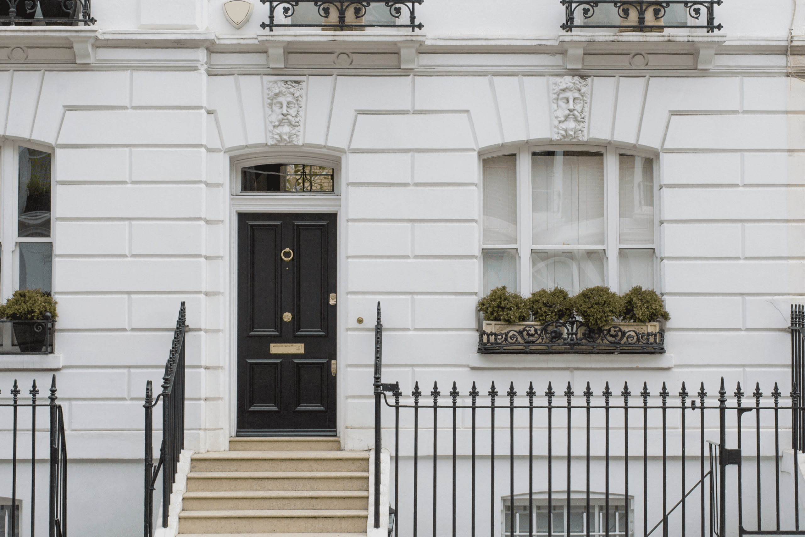 The image shows the front of a classic white townhouse with a black door, flanked by two windows with decorative railings and potted plants. A set of steps leads up to the entrance, and a black iron fence lines the foreground.
